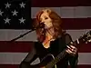 A woman with red hair wearing black clothing and hoop earrings playing the guitar and singing into a microphone. The American flag appears in the background.