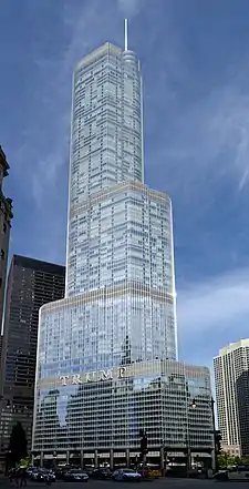 A tall silver skyscraper sits at a jog in the river beyond a bridge. The river and other along its banks buildings are in the foreground.