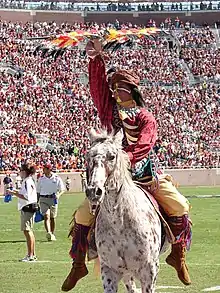 A brown and white spotted horse ridden by a sports mascot in modern-day Native American attire waving a flag stands on a sports field. More people are visible on the field, and a large crowd fills the stadium seating in the background.