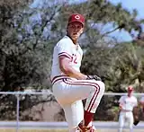 A man wearing a white baseball uniform and a red cap with a white "C" on it winds up to throw a baseball from the pitcher's mound