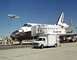 駐機中のエンデバー号。上空にはシャトル輸送機で空輸されるコロンビア号が見える