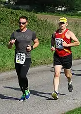 Two persons exercising on a paved road in a park. The weather is sunny.