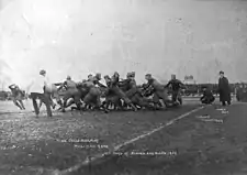 A grainy black and white photo of teams in sports jerseys and light padding contesting a play near the touchline, while watched by officials, coaches and crowds.