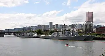 ポートランド市内を流れるウィラメット川の右岸から対岸を眺める。岸辺にはアメリカ海軍の駆逐艦が数隻停泊し、その隙間から川岸が公園になっているのが見える。公園の奥にはダウンタウンのビルが立ち並び、その向こうに山並が見えている。空は晴れているが雲が多い。