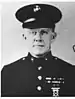 Head and shoulders of a white man wearing a peaked cap and a dark military jacket with ribbon bars on the left breast and bright buttons down the center.