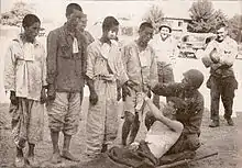 ひとりの兵士に支えられたシャツを着ていない白人男性が、4人並んだアジア系男性のひとりを指差す