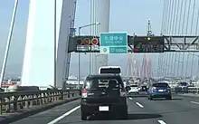 画像左:第二東名と伊勢湾岸道路の境界に設置された可変式速度規制標識と「ここから一般有料道路」の標識板（下り東海IC付近、KP30+649）。トレーラーの車線指定標識も設置された。画像右:可変式速度規制標識は平均1.3 km毎に設置されている。画像は高速車50 km/h規制時で、最低速度標識は消灯している。