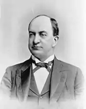 A balding man with dark hair wearing a black jacket and tie and white shirt.