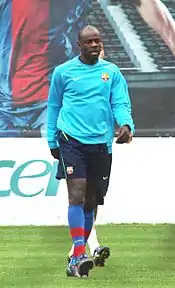 A dark-skinned man wearing a teal shirt, black pants, and blue, deep red socks.