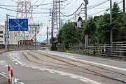安全地帯の標識や安全防護設備の柵も無く、道路標示の白線が引かれ青色で囲むように塗りつぶしただけの停留場。（万葉線新吉久停留場）