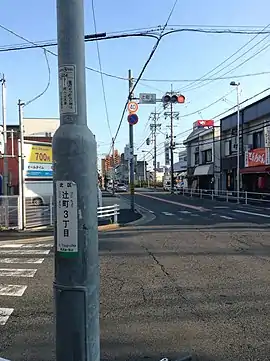 辻町の読みの揺れ(奥の交差点はローマ字で「つじまち」と表記され、手前の行政設置のステッカーには「つじちょう(つじまち)」と併記されている。)
