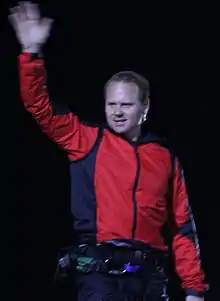 A man in a red jacket waves with his right hand raised high in the air