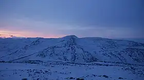 雪に覆われた山。視界の全体が暗い青色に見え、左側からは暗いピンク色の輝きが地平線から立ち上っている。これでも太陽の正中時の風景である。