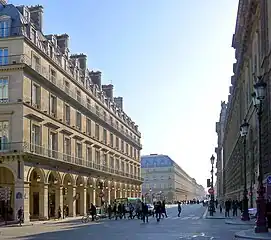 ルーヴル宮殿からパレ・ロワイヤル広場近くリヴォリ通りを見る (La rue de Rivoli au niveau de la place du Palais-Royal, devant le palais du Louvre.)。(パレ・ロワイヤル広場との角に建つ) 向かい建物は大型骨董店ルーヴル・デ・アンティケール (fr, 1978年9月開業)。