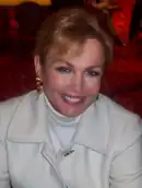 Portrait photo of a woman with blonde hair in her late 50s smiling at the camera. She is wearing a white coat and white turtleneck underneath.