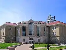 A large, brown building with towering windows, a clock tower, and a statue of a man in front