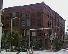 A large 4-story red brick building with many windows. The building is on a street corner with the front and one of the sides visible.