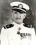 Head and shoulders of a white man with a thin, upturned mustache wearing a white peaked cap with black visor and a white jacket with dark shoulder boards, a row of medals on the left breast, and a star–shaped medal hanging from the neck.