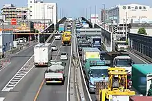 画像左 : 名古屋港で船からトラックに積み替えられたコンテナは中部経済を支えるために各地へ輸送されるが、名四国道の渋滞によって物流が滞る問題があった。伊勢湾岸道路はその切り札と目された（飛島ふ頭、旧西4区のコンテナターミナル）。画像右 : 日中の名四国道（名古屋市港区品川町付近）。1980年代半ばには既に物流動脈としての限界を露呈し、危機感が高まっていた。