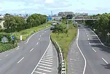 画像左:名二環着工前の富田IC付近(名古屋市中川区かの里付近)。専用部の用地は一般部中央に確保済であった。画像右:工事中の名古屋西JCT - 飛島JCT間(国道23号交差点付近 2017年5月)。港湾物流に関わる大型車の渋滞が著しいことから専用部の開通が期待された。