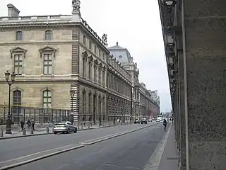 リヴォリ通りから見るルーヴル宮殿 (Le palais du Louvre, vu d’entre deux arcades de la rue de Rivoli.)。撮影地点の同アーケード建物は、大型骨董店ルーヴル・デ・アンティケールが入る、サントノレとリヴォリ通りとに挟まれた同大型区画上の建築物。元々は、旧ルーヴル百貨店 (fr) が1855-1974年にかけてあった地。ゾラ作『ボヌール・デ・ダム百貨店』(1883年) も参照。