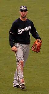 A man wearing a navy blue Brewers jersey, gray pants, navy blue cap, and outfielder's glove on his left hand walking in the outfield.