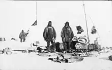  On the left is a snow cairn with flags. Three men are nearby, and assorted equipment is strewn on the snow.