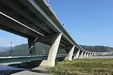 画像左 : 新安倍川橋と葵大橋。長さ1.7 kmのこの橋は県道と第二東名の二層構造で、画像奥の川岸に新静岡ICが敷設されている。インターを市街地から大きく外れた場所に造ったために新たな道路が必要になり、道路が増殖を続ける構図であるとして批判された。別のマスコミはこの橋を皮肉を込めて「土木技術の展覧会の面もある」と書いた。画像右 : 静岡市清水区和田島に架かる興津川橋。200以上の橋梁建設に関わってきた建設作業員は、山の中でこうした大きい橋を造るのは初めてで驚いたという。この橋の一山向こうに掘削された清水第三トンネルは公団が「第二東名の技術の結晶」と呼び、このトンネルの嘱託実験を請け負った大学教授が「唯一残った大規模な実験場だ」というインタビュー内容を皮肉交じりに記事化した。
