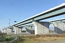 整備計画区間における起点の海老名南JCT付近。画像左 : 住宅街に囲まれた更地である東京方面の本線建設用地最東端からジャンクションを望む。これより東（画像手前）の区間は基本計画以下にとどまっている。画像右 : 海老名南JCTに連結する相模川橋。橋脚は将来の拡幅が困難であることから完成形の片側3車線とランプ部を含めた仕様であるが、その先の横浜、東京方面に延伸する目処は立っていない。