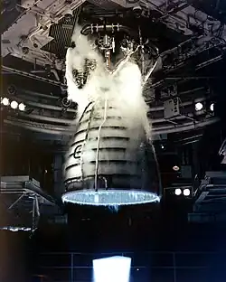 噴射しているロケットエンジン。青い炎が周囲に複数のパイプがある釣鐘状ノズルから噴き出している。ノズル最上部は蒸気で覆われていて天井から吊り下げられている複雑な配管部に取り付けられている。後ろにはトランジェント装置の一部が見える。
