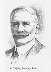  A well-dressed man with greying hair and a large moustache stands looking directly at the camera. In his right hand is a cigar.