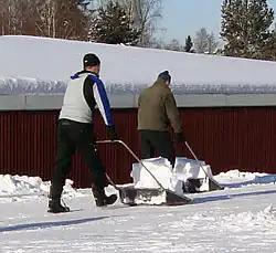 平らな積雪面においてスノーダンプを使用して雪を運搬する2人の人物の後ろ姿。
