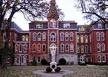 A statue sits in front of a large 4 story red brick building. To the right of the entrance, the building goes in a little ways, and then back out. To the left of the entrance is a small 2 story area connecting the main part of the building to the rest of it. Above and behind the 2 story part is a 4 story part of the building