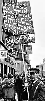 A man in coat, tie, glasses, and a baseball-like cap carries a tall sign in a crowded street. The sign says "LESS LUST, BY LESS PROTEIN: MEAT FISH BIRD; EGG CHEESE; PEAS BEANS; NUTS." Then "AND SITTING, and in smaller letters below that "PROTEIN WISDOM".