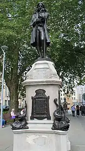 Larger than lifesize bronze statue of man in period clothes, standing with one hand on a staff, the other raised to his chin. It is on a white stone pedestal with inscription Edward Colston Born 1636 Died 1721", and bronze inscribed plaques below. Large bronze dolphins are on each corner of the base. It is in an urban setting with a large tree behind and above it.