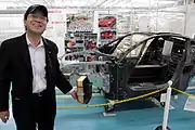 見学者にCFRPボディのカットモデルを解説する棚橋晴彦。後ろに置かれているのは製造途中の370号車（元町工場）