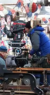 大型の映画用カメラを覗き込む、青いジャケットと黒い帽子の男性。頭上には赤白青の幔幕が飾られている。