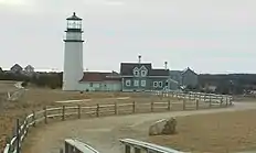 The lighthouse is in the background of the photo, and in the foreground is a large rock, or boulder. In the far distance, on the right, is the Pilgrim Monument tower in Provincetown.