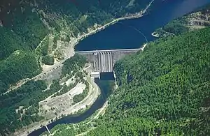 画面の中央にダムの堤体が捉えられた。航空写真。ダムの貯水湖が画面の右上に向けて広がっており、ダムの下流は左下へ延びている。川は森におおわれた斜面の間を流れており、右岸側が切り立っていて所どころ岩盤がのぞいており、湖面沿いから堰堤の横を通るように道路が延びている。堤体直下の右岸には発電所の建物が見える。