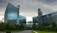 Two geometric all glass towers connected by a central atrium stand in front of a grassy walkway and under a dark and cloudy sky