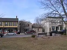 A row of 3 older buildings lines the far side of a street. On the near side is grass from a park with two park benches