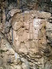 写真は彫刻された岩を示している。濃い灰色の縞模様が陥入した青灰色の岩の上の乳灰色の岩に彫刻が現されている。ヒッタイト王が左に描かれており、彼はひげを生やし、2本のブドウの枝を持っている。彼に面して描かれている神は、経時的な荒廃のため、頭と足の部分が失われている。