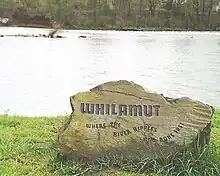 ウィラメット川の芝生に覆われた岸辺に "Whilamut"と刻まれた巨石があり、その向こうに川面が広がり、対岸には森が広がっている。