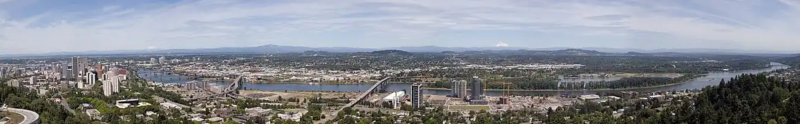 ポートランド市街の南に位置するウィラメット川左岸の丘陵から東を望む。画面の右からウィラメット川が流れてきて画面の右半分に広がるポートランド市街地を縫って流れていく。画面の中央にロス・アイランド橋がありそのすぐ右にロス島が見える。そこから下流に向けていくつもの橋がポートランド市街地を結んでいる。画面奥の地平線には山頂が雪に覆われた山が三峰写っている。