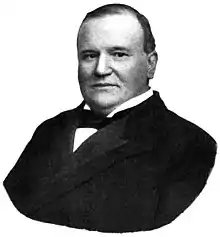 A heavy-set, balding man in his fifties wearing a black coat and tie and a white shirt, facing left